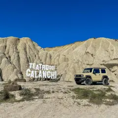 Un fuoristrada beige parcheggiato davanti al **teatro dei calanchi**, un paesaggio lunare con montagne erose e un cartello in legno.