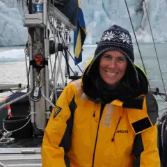 Glad kvinna i gul jacka på segelbåt framför glaciär. Drömmen att segla jorden runt är nära.