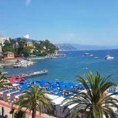 Plage animée à Santa Margherita Ligure, Italie, avec parasols bleus, cabines rayées, palmiers et bateaux sur la mer bleue.