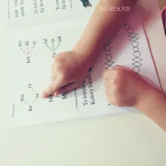 Dziecko ćwiczy czytanie, wskazując palcem na litery alfabetu i sylaby w książce. Obok widnieje wzór z figur geometrycznych.