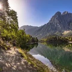 Senderista camina por el sendero junto al estany de Basturs, reflejando la majestuosa montaña y el bosque en sus aguas tranquilas.