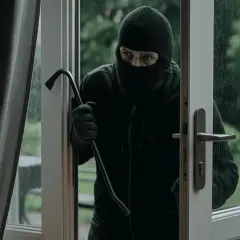 Un cambrioleur masqué, prêt à forcer une porte avec un pied-de-biche, illustre le danger du home jacking et l'importance de la protection.