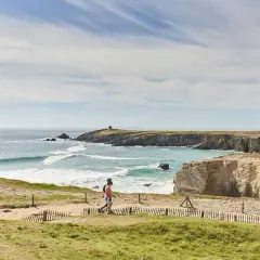 Deux personnes marchent sur un sentier côtier, découvrant les plus beaux endroits de Quiberon. Falaises dorées, mer turquoise et ciel bleu.