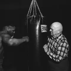 Mike Tyson trains with a heavy bag, his muscular physique evident as he prepares for a punch. An older man in a plaid shirt watches intently.