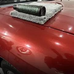 Butelka wosku samochodowego SWAG na szarej mikrofibrze leżącej na błyszczącym, czerwonym lakierze auta.