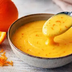 Une cuillère dégouline d'une sauce maltaise onctueuse, parsemée de zestes d'orange. Une orange entière et une tranche sont à proximité.