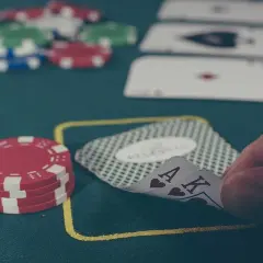 Dłoń trzyma karty do gry (As i Król) na zielonym stole do pokera, obok stosu żetonów.