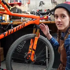 A woman in a blue plaid shirt and beanie explains bike maintenance, pointing to the orange frame and front suspension of a mountain bike.
