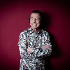 Raphael, sonriente y con los brazos cruzados, posa ante un fondo rojo. Su camisa estampada evoca la energía de sus conciertos.