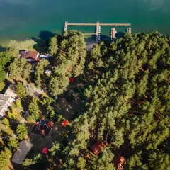 Malowniczy ośrodek nad jeziorem otoczony lasem. Widoczne budynki, pomost z łódkami i plac zabaw.