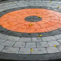 Kreisrunder Weg aus grau und orangefarbenen Pflastersteinen, die wie ein Ziel aussehen. Einige gelbe Blätter liegen darauf.