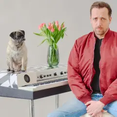Ein Mops sitzt auf einem Keyboard, daneben ein Mann in roter Jacke. Eine Vase mit Tulpen steht zwischen ihnen. Eine gute Nachricht: Keine Gefahr!