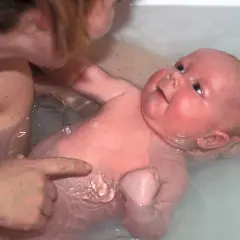 Ein Baby genießt sein Bad, während eine Hand es sanft hält und eine andere auf seine Haut zeigt.