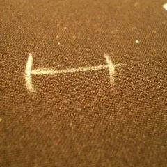 Na brązowym materiale zaznaczono białą kredą kształt przypominający literę "H", przygotowanie do ręcznego obszywania dziurek.