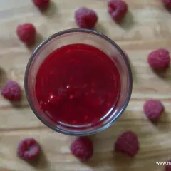 Szklanka z malinową frużeliną otoczona świeżymi malinami na drewnianym tle.