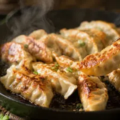 Gorące, rumiane pierożki gyoza parują na patelni. Jedna sztuka jest podnoszona pałeczkami, gotowa do zanurzenia w sosie.