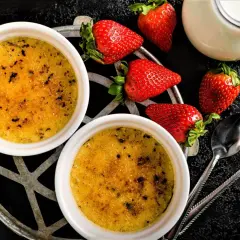 Zwei cremige Crème brûlée mit karamellisierter Kruste, garniert mit frischen Erdbeeren und einem Milchkännchen.