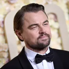 Leonardo DiCaprio wiek: aktor w czarnym smokingu z muszką, z siwiejącymi włosami i zarostem, na tle kwiatowej dekoracji.