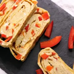 Roladki omlet tamago z papryką, pokrojone na plastry, prezentują się apetycznie na czarnej desce.