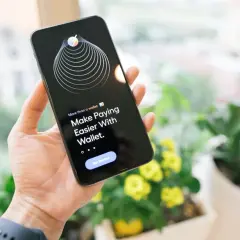Dłoń trzyma iPhone'a z ekranem wyświetlającym aplikację Wallet i napis "Make Paying Easier With Wallet".