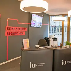 Hier ist die Rezeption der IU Internationale Hochschule. Ein Bildschirm zeigt Bilder, und ein Plan weist den Weg. Was ist die IU? Eine Hochschule, die deine Zukunft beginnt.