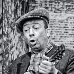 Stanisław Bard Starej Warszawy, w czapce i z gitarą, śpiewa na tle ceglanej ściany.