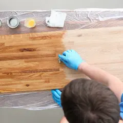 Mano con guanto blu applica una finitura su legno, creando una patina. Diverse lattine di vernice e un panno sono visibili.