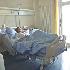 Młoda kobieta leży w szpitalnym łóżku, otulona niebieskim kocem. Jej hospitalizacja przebiega spokojnie, a przez okno wpada jasne światło.