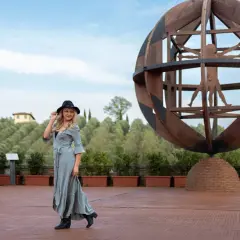 Donna con cappello nero e abito lungo in un'area panoramica del vinci centro storico, con la scultura dell'Uomo Vitruviano sullo sfondo.