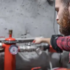 Uomo con barba controlla un manometro su un tubo rosso. Sento rumore nei tubi dell'acqua, forse un problema da risolvere.