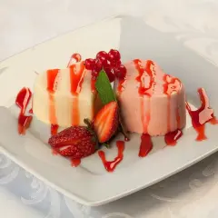Dwa sercowate desery pana cotta, jeden waniliowy, drugi truskawkowy, polane sosem truskawkowym, ozdobione świeżymi truskawkami i porzeczkami.