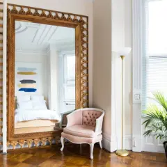 A grand pier mirror with an ornate gold frame reflects a bed and abstract art. A pink velvet chair sits beside it.