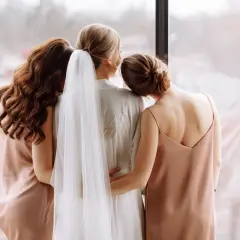Trzy kobiety w jedwabnych szatach stoją przy oknie. Jedna z nich, panna młoda, ma na sobie białą szatę z welonem. Jej druhna obejmuje ją czule.