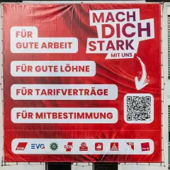 Großes rotes Banner mit der Aufschrift "Mach dich stark mit uns". Es wirbt für gute Arbeit, Löhne, Tarifverträge und Mitbestimmung. Was ist ver.di? Die Antwort steht unten.