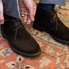 Mani che allacciano stivaletti scamosciati marroni, un dettaglio di stile per polacchi uomo eleganti su un tappeto persiano.