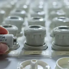 Dłoń trzyma bezpiecznik topikowy 25A 500V E, na tle rzędu innych bezpieczników.