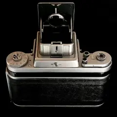 A vintage Pentacon Six camera, with its viewfinder open, sits on a reflective black surface.