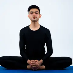 Młody mężczyzna w czarnym stroju wykonuje pozycję baddha konasana na niebieskiej macie.