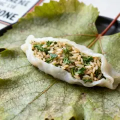 Une feuille de vigne farcie, garnie de riz et d'herbes fraîches, repose sur une grande feuille de vigne verte sur une assiette noire.