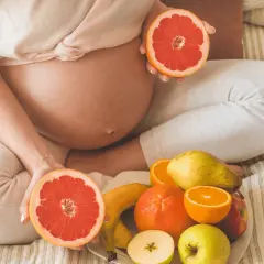 Ciężarna kobieta trzyma połówki grejpfruta, obok talerz z owocami. Czy w ciąży można jeść pomarańcze? Tak, to zdrowe!