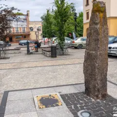 Kamienny pomnik i tablica z napisem "Kamienna" to ciekawe atrakcje na rynku. Obok ławki i drzewa, w tle budynki i samochody.