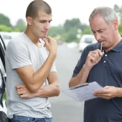 Młody mężczyzna zamyślony obok samochodu, instruktor z notatkami i długopisem przy ustach, przygotowują się do egzaminu na prawo jazdy B.