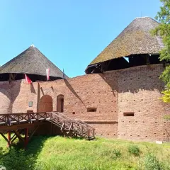 Zamek międzyrzecz z drewnianym mostem i wieżami o stożkowych dachach. Czerwona cegła i zieleń otoczenia.