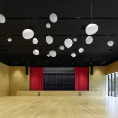 Moderne Aula mit Holzwänden, Bühne mit roten Vorhängen und vielen weißen Hängelampen. Die nobelgusch pfedelbach ist bereit für Veranstaltungen.