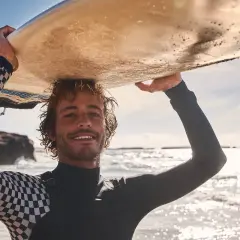 Lächelnder Surfer mit lockiger surfer frisur trägt Neoprenanzug und hält Surfbrett über dem Kopf.