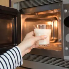 Ręka wkłada szklankę mleka do mikrofalówki. Czy podgrzewanie mleka w mikrofali jest szkodliwe? To pytanie nurtuje wiele osób.