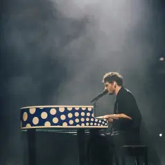 Dawid Podsiadło na koncercie, grając na pianinie w niebiesko-białe kropki, otoczony mgłą sceniczną.