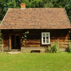 Drewniana chata z glinianym dachem, typowa dla skansenów w Polsce, otoczona zielenią i drewnianym płotem.