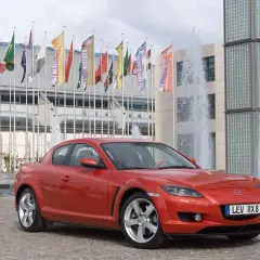 Czerwona Mazda RX-8 zaparkowana przed budynkiem z fontannami i flagami.