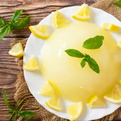 Un rinfrescante gelo di limone, guarnito con spicchi di limone e foglie di menta, su un piatto bianco, circondato da limoni e menta su un tavolo di legno.